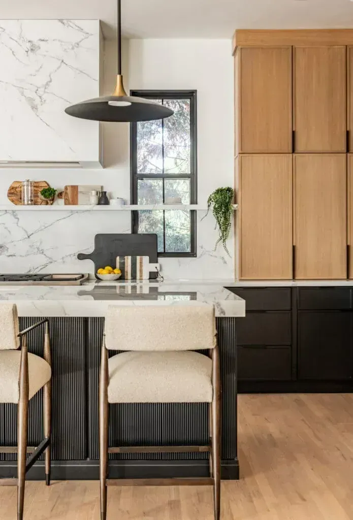 Modern kitchen with marble countertops, black island, light wood cabinets, and black window.