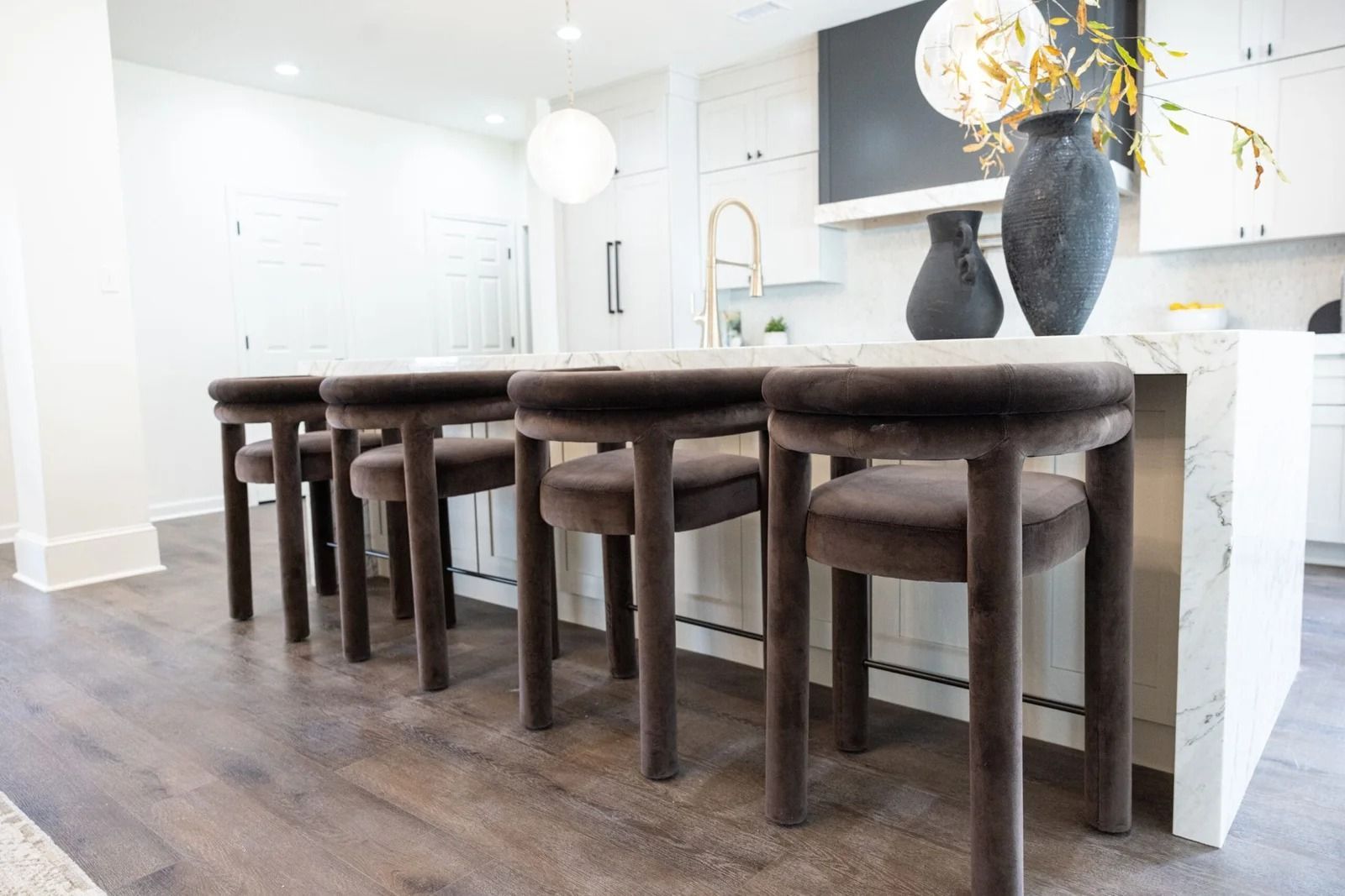 Kitchen island with five velvet-covered bar stools. Dark wood floors, white cabinets, and decorative vases.