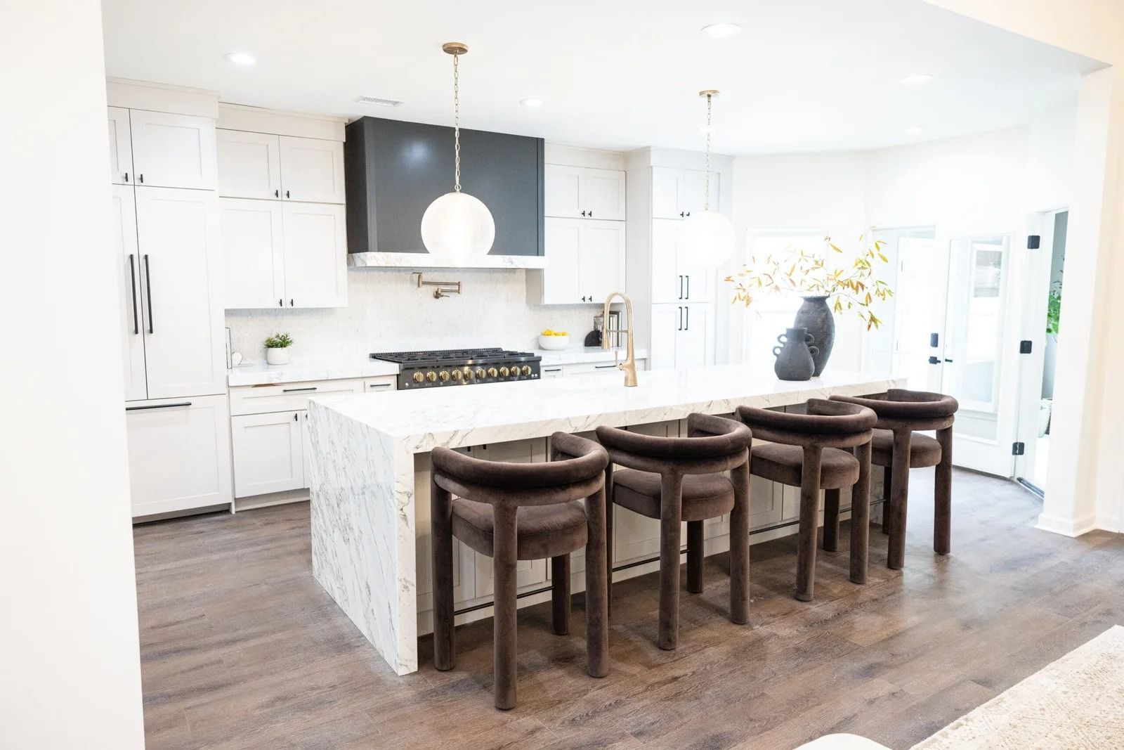 Modern white kitchen with island seating, dark hood, and brown stools.