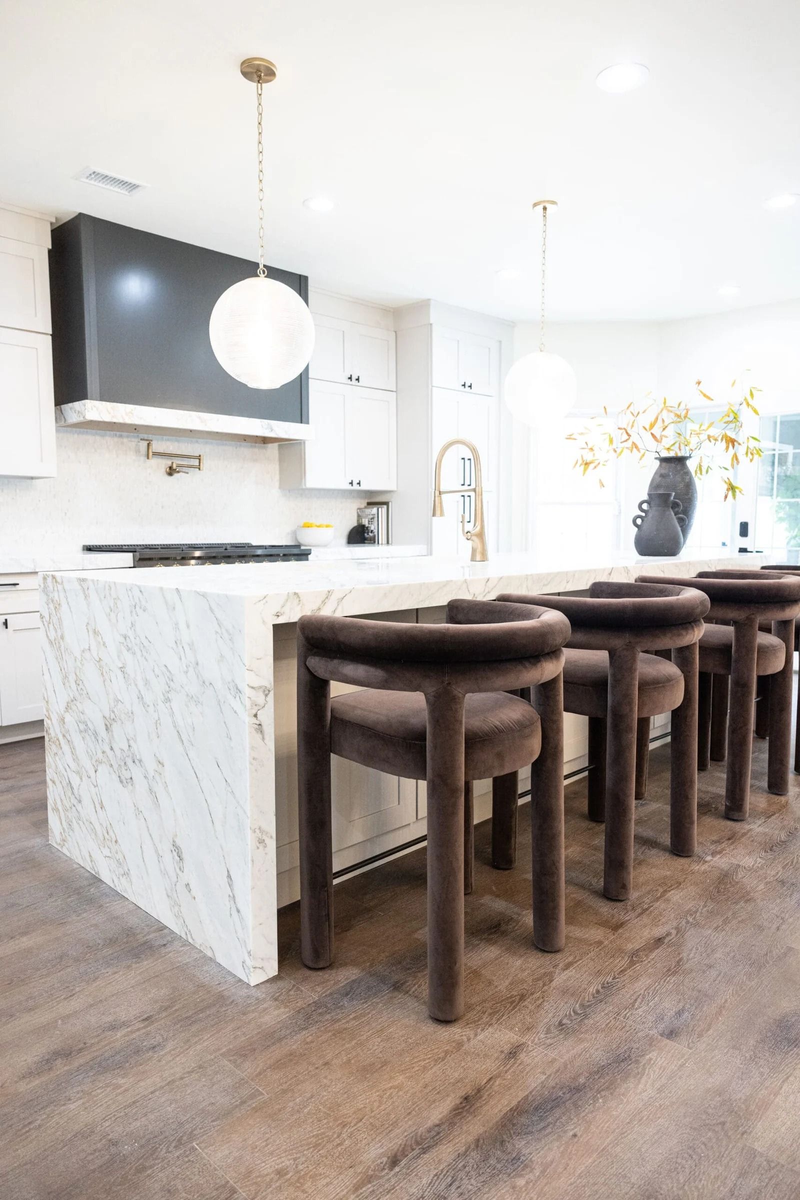 Modern kitchen with marble island, dark brown bar stools, white cabinets, and gold light fixtures.