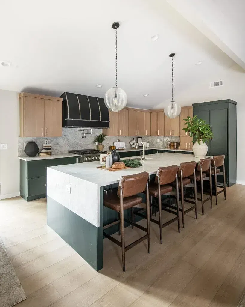 Modern kitchen with two-tone green and wood cabinets, white countertops, and leather bar stools at the island.