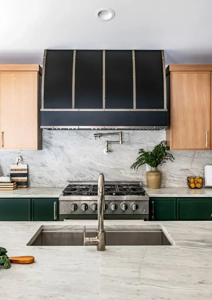 Modern kitchen with stainless steel appliances, dark range hood, green and light wood cabinets, and marble countertops.