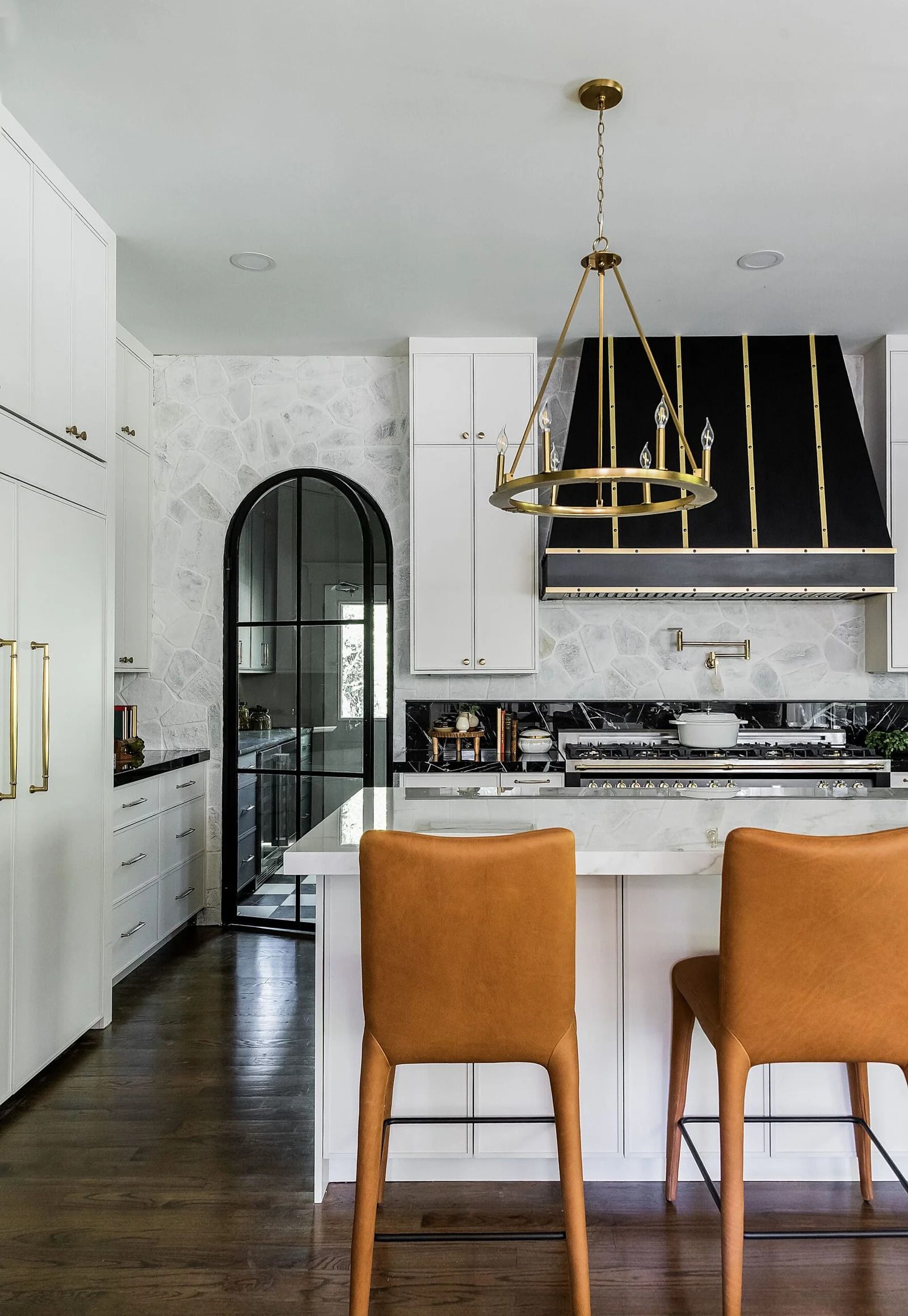 A kitchen with white cabinets, marble backsplash, a black and gold range hood, and two brown leather bar stools.