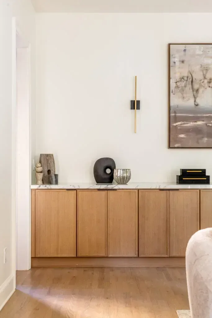 Wooden cabinets with marble countertop and decorative items against a white wall with artwork and a sconce.