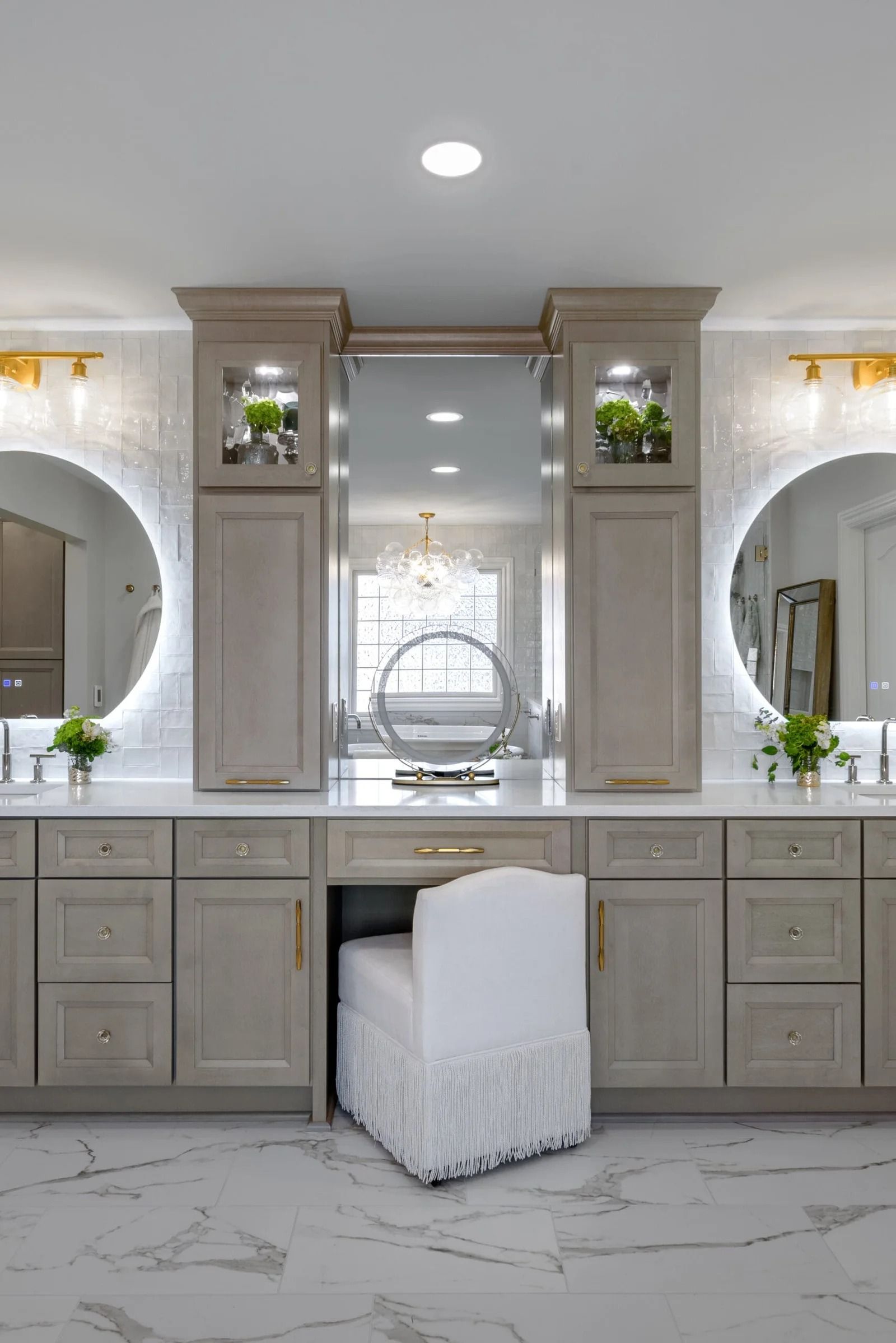 Bathroom with a vanity and a makeup chair. Gray cabinets, round mirrors, and white marble floor.