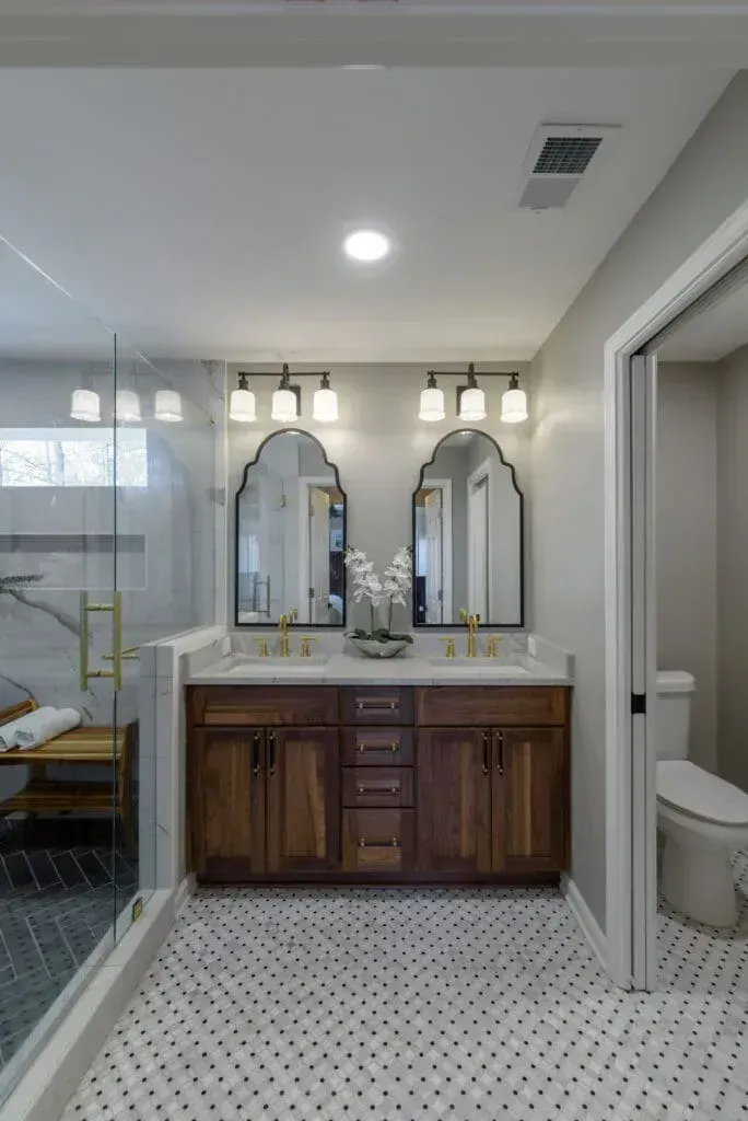 Bathroom with wood vanity, arched mirrors, and glass shower. Black and white tiled floor.