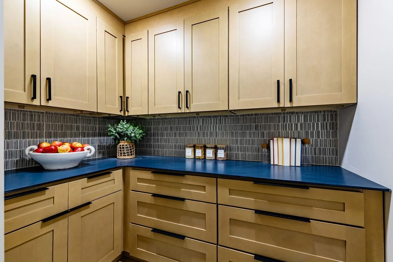 A pantry with light wood cabinets, dark blue countertop, and a tiled backsplash.