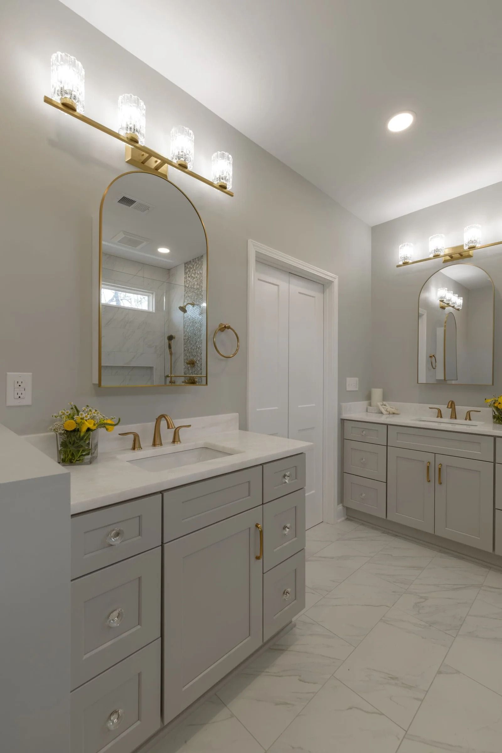 Two bathroom vanities with mirrors, lights, and gold fixtures in a light gray and white bathroom.