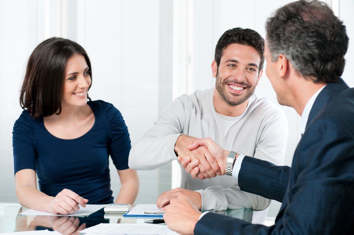 Three colleagues shaking hands at a conference table, with a smiling woman seated beside them.