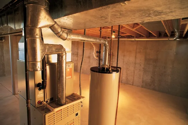 Furnace and water heater in a basement with silver ductwork, concrete walls, and wood beams.