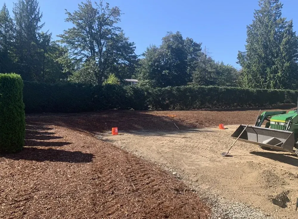 A construction site with a tractor. Mulch and gravel areas with trees in the background.