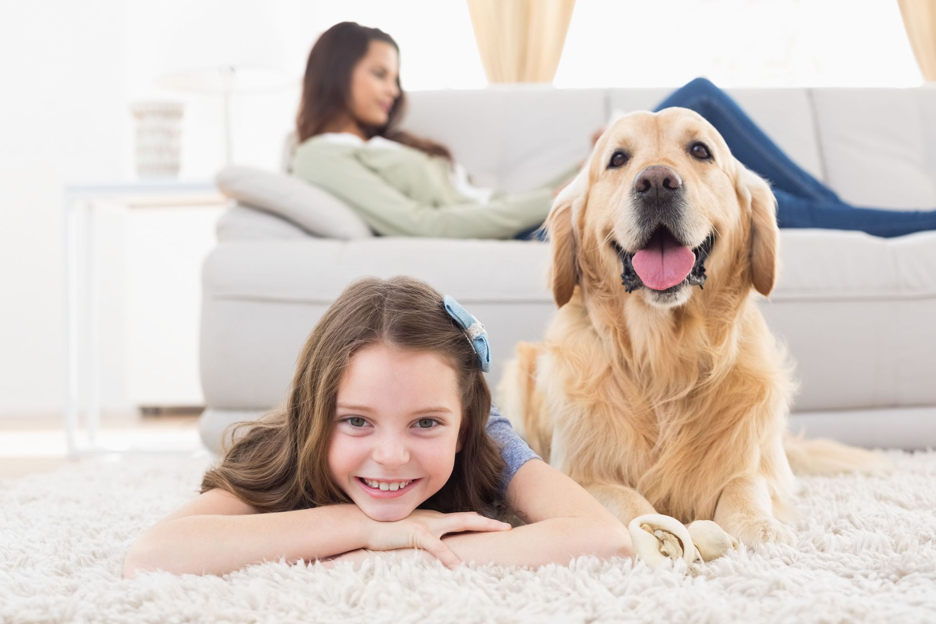 Girl smiling with a Golden Retriever on a carpet, woman on couch in background.