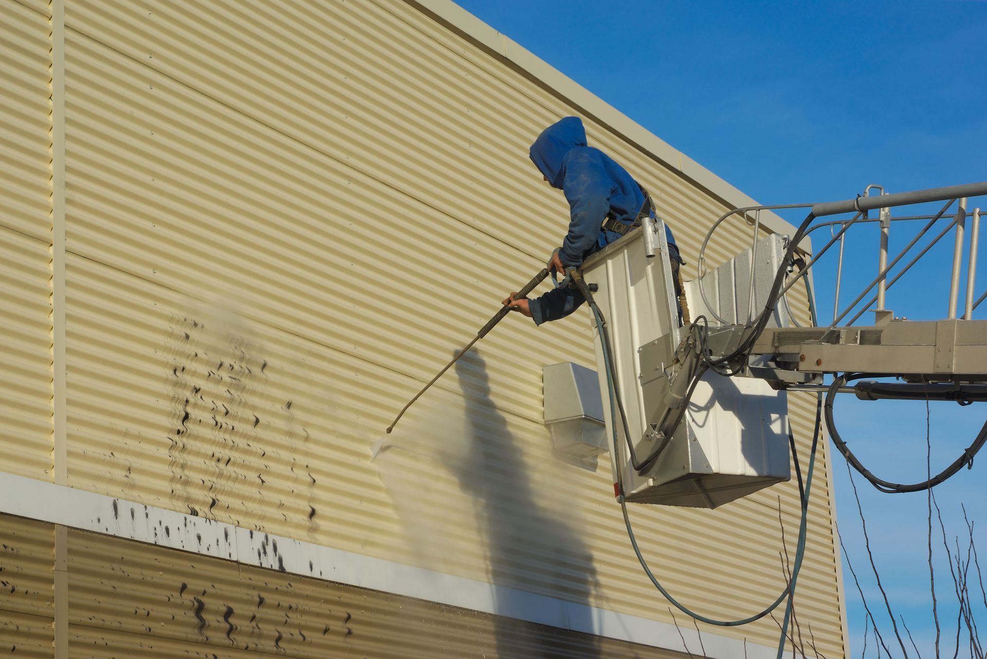 Worker in blue rain gear pressure-washing a beige building from a lift basket.