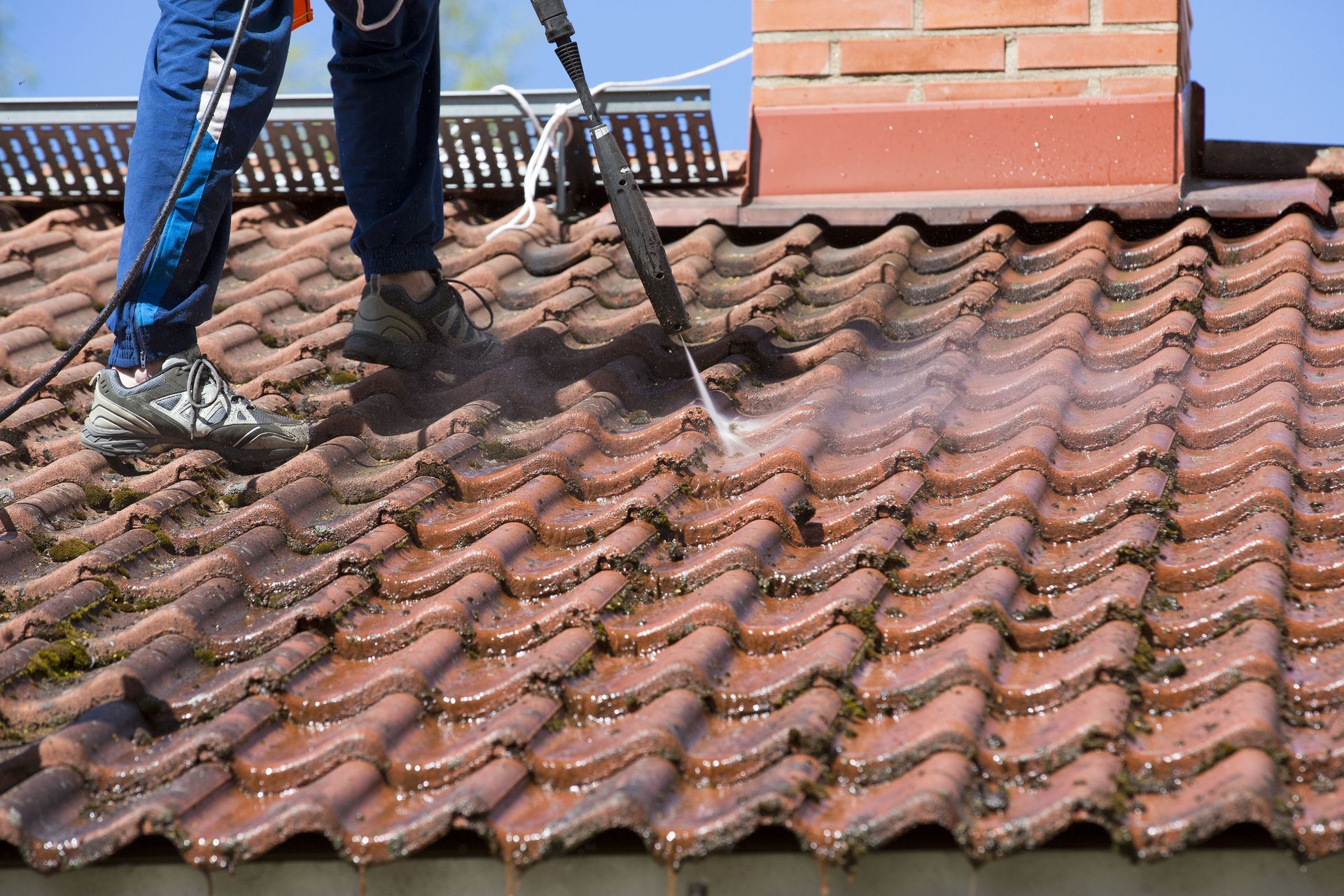 Worker cleaning a mossy terracotta tiled roof beside a brick chimney