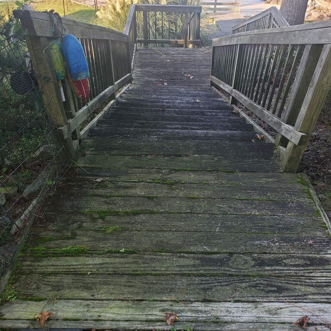 Weathered wooden boardwalk with railings leading downhill toward water, with a person in a blue jacket on the left.