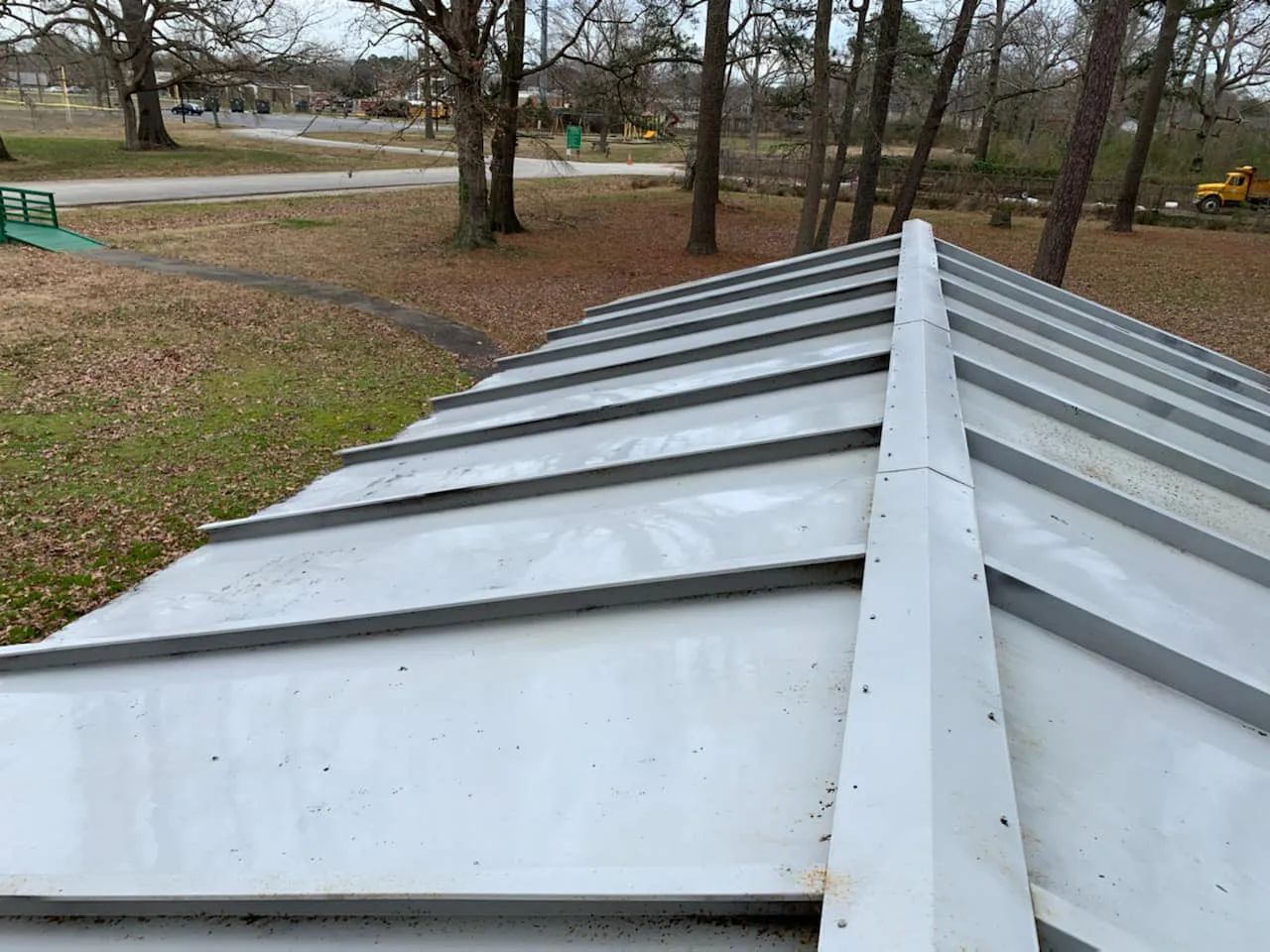 Silver metal roof with central ridge, viewed from above in a leafless park near a road.