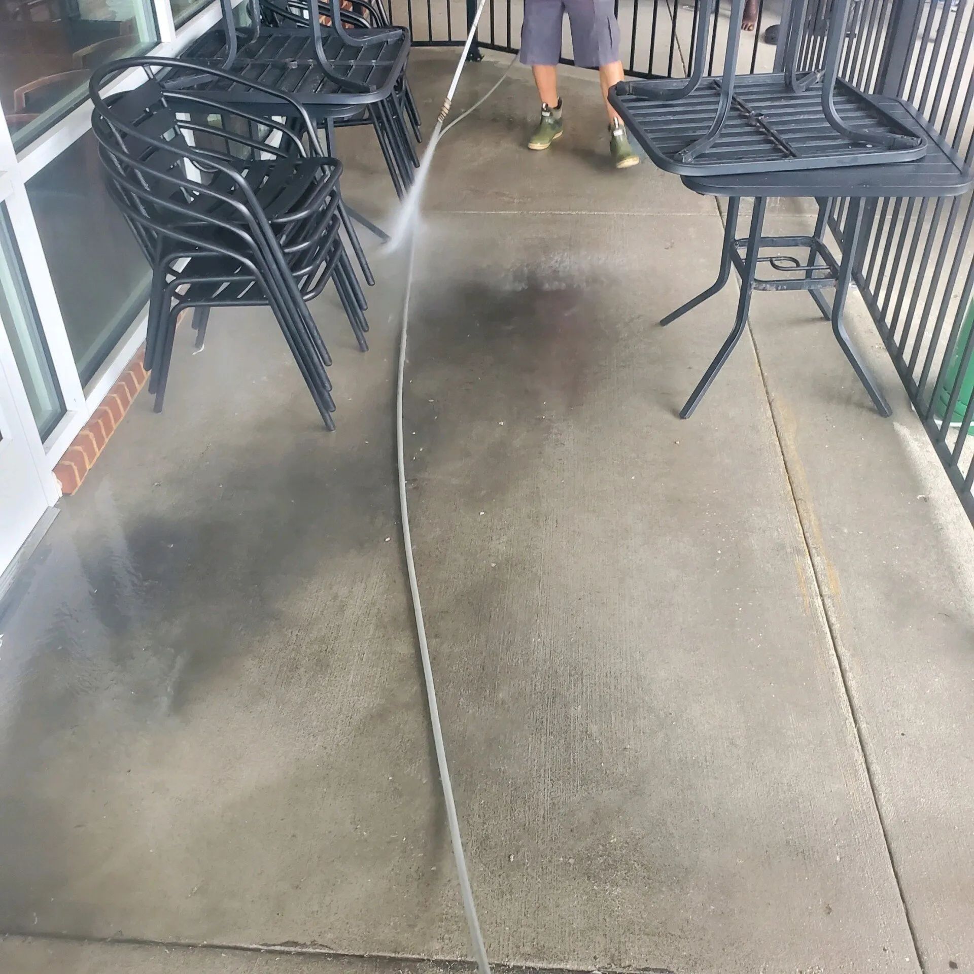 Outdoor café walkway with stacked black chairs and tables on a concrete patio, with a person standing in the distance