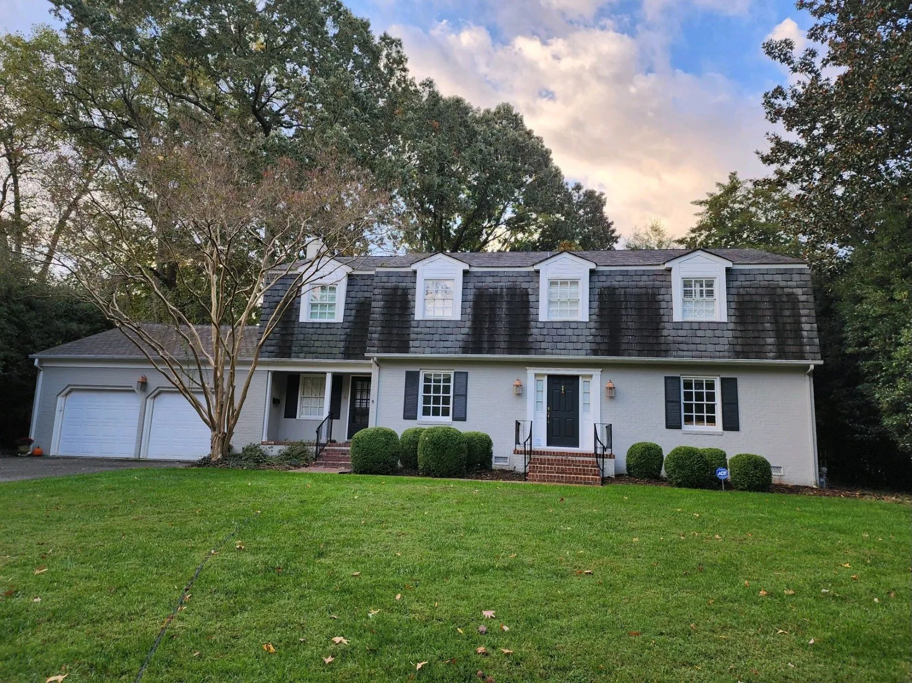 White two-story house with dark shutters, garage, and a large green lawn under a cloudy sky
