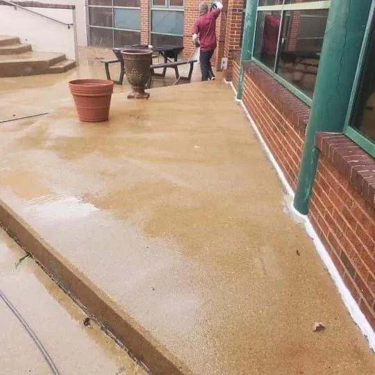 Person sweeping a wet patio beside a brick building, with a large planter and benches nearby.