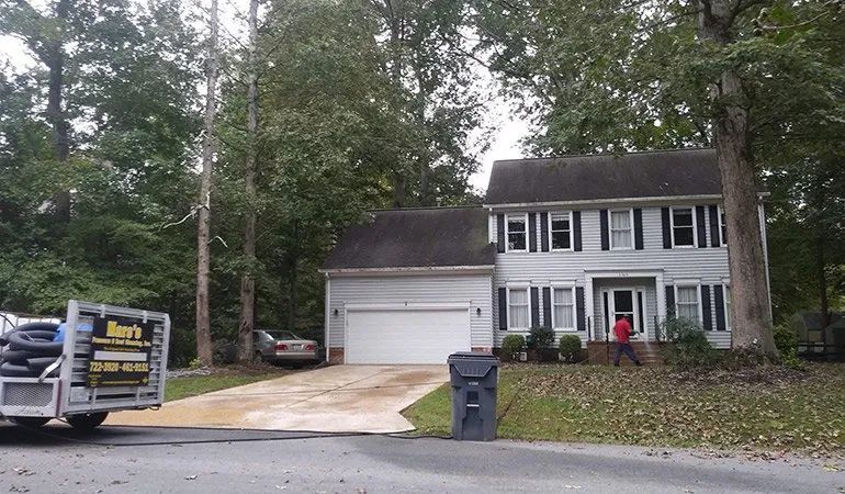 Suburban two-story white house with attached garage and driveway, surrounded by trees and a for-sale sign.