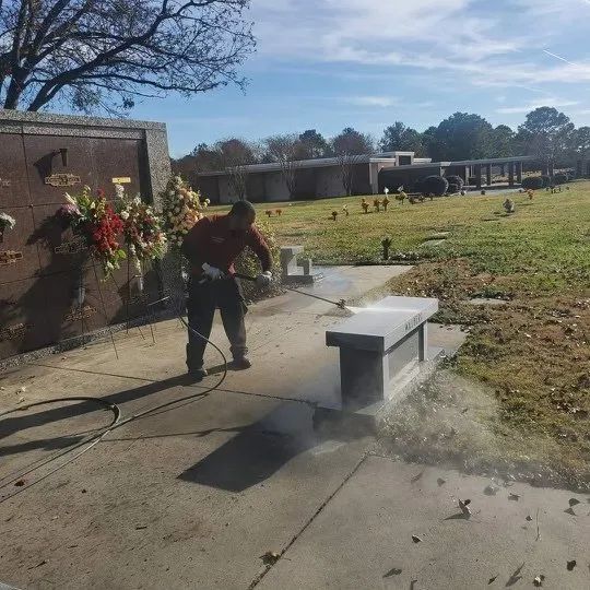 Person pressure-washing a white bench on a sunny sidewalk beside a grassy field and building