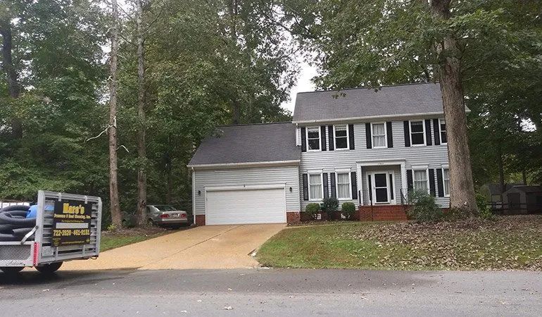 Suburban two-story house with white siding, black shutters, attached garage, and a real estate sign by the driveway