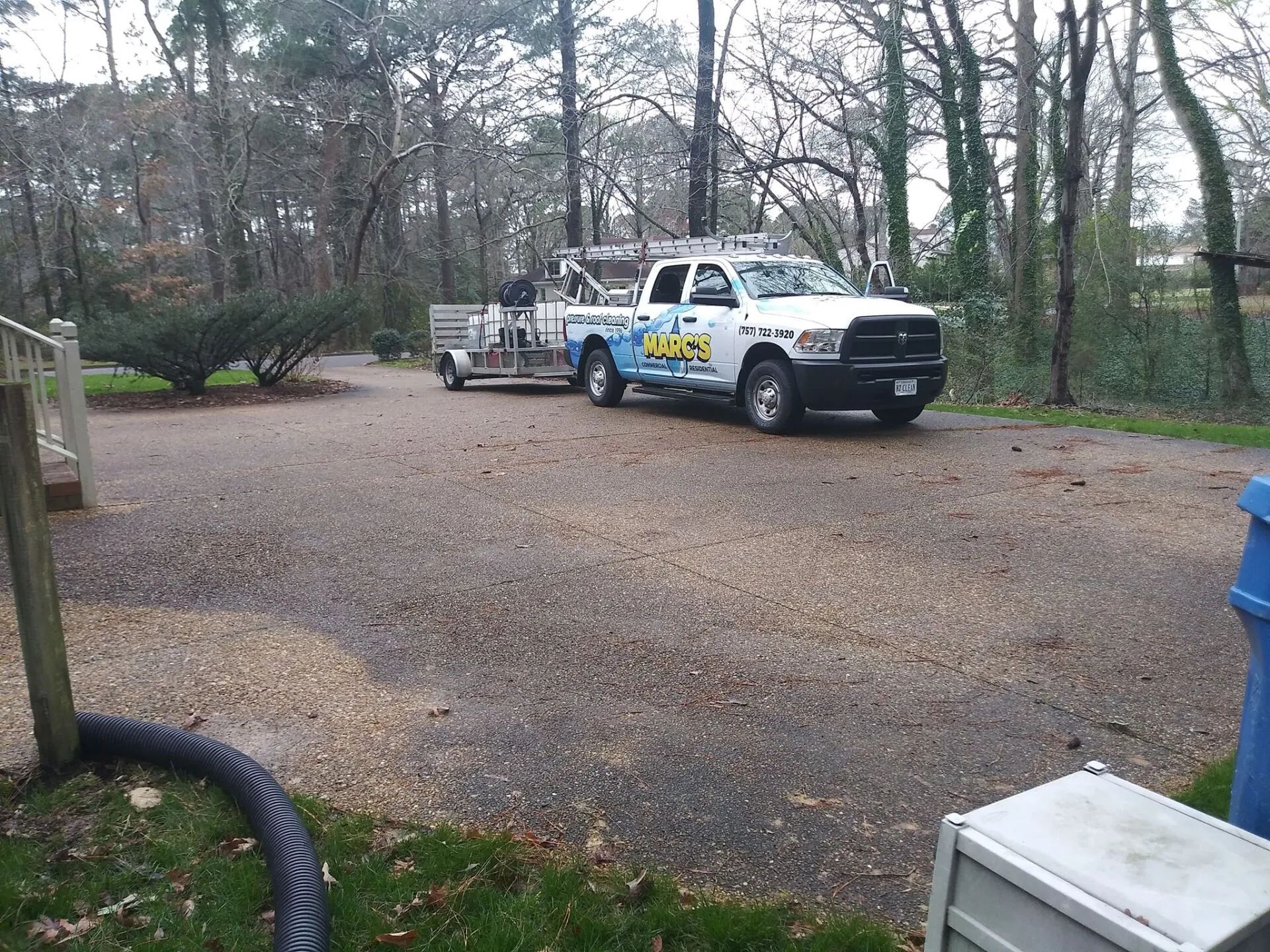 White tow truck in a gravel driveway near trees, towing a small trailer in a wooded yard.