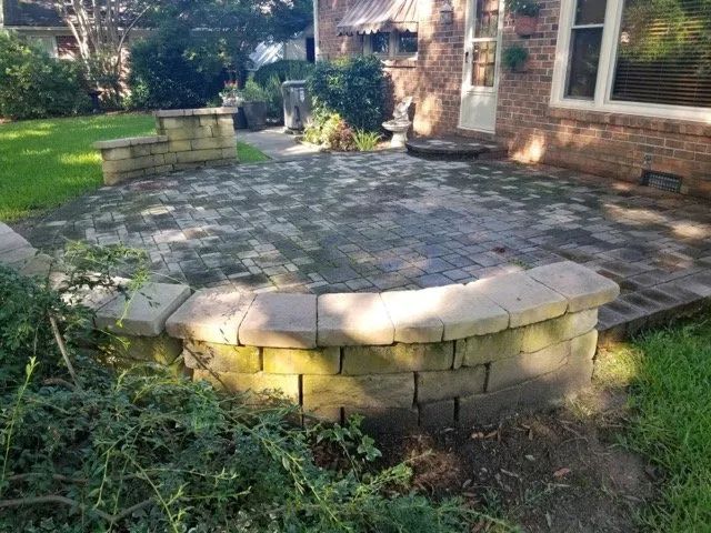 Backyard stone patio beside a brick house, bordered by low retaining walls and surrounded by grass.