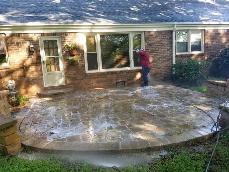 Person spraying water on a wet stone patio beside a brick house with sliding glass doors.
