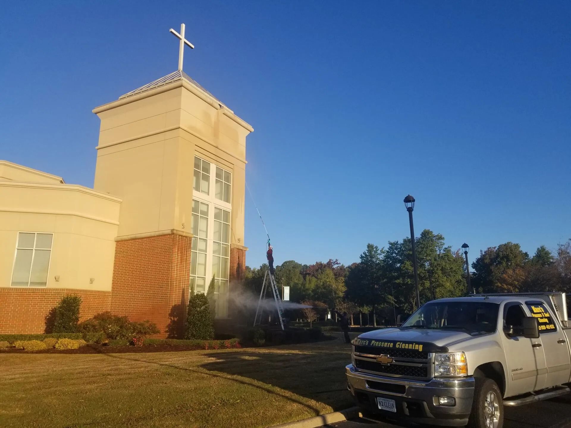 White church tower with cross and fountain, with a pickup truck parked beside it under a blue sky.