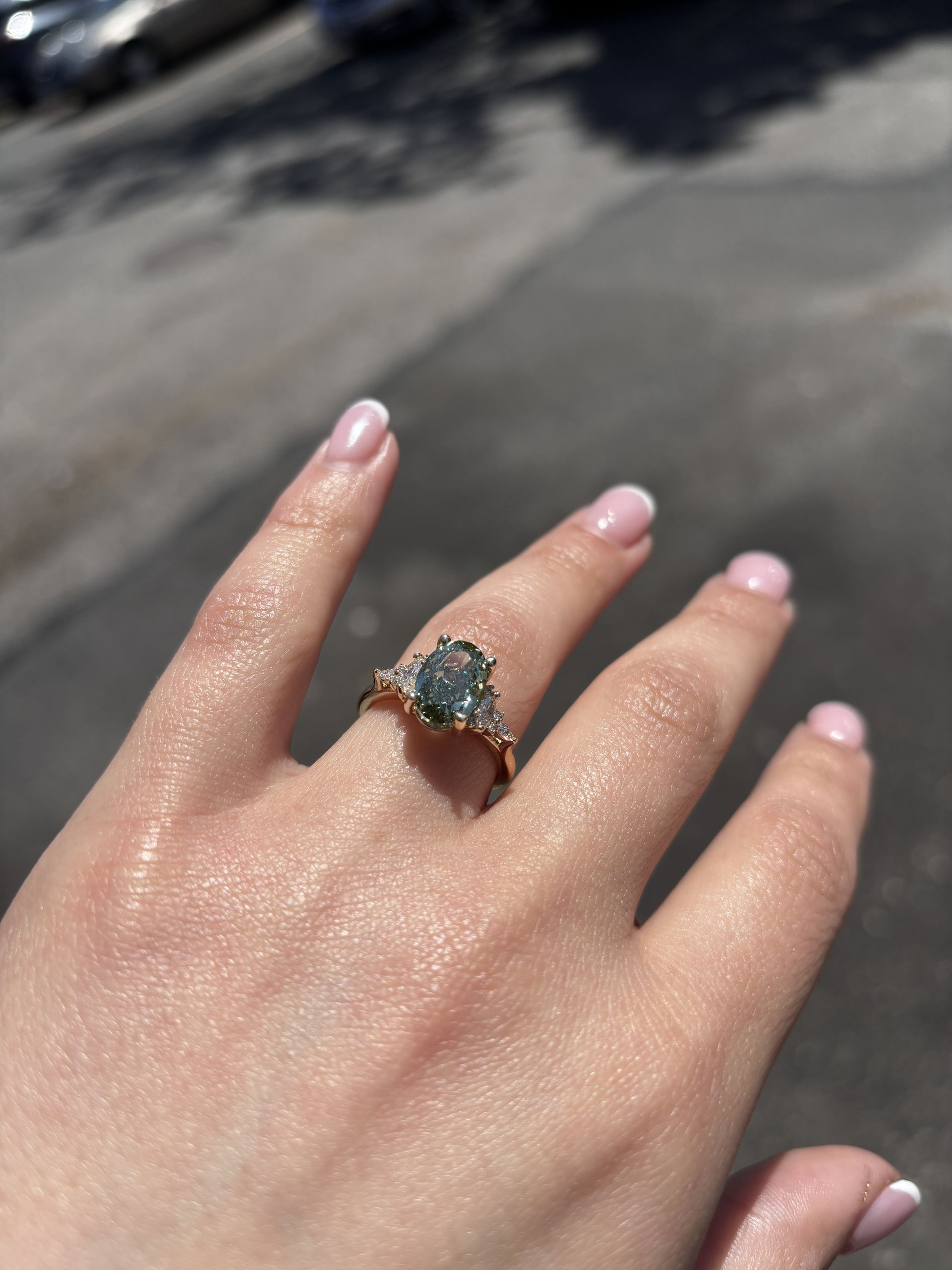 Hand wearing a gold ring with a green stone, held up in sunlight against a blurred pavement background