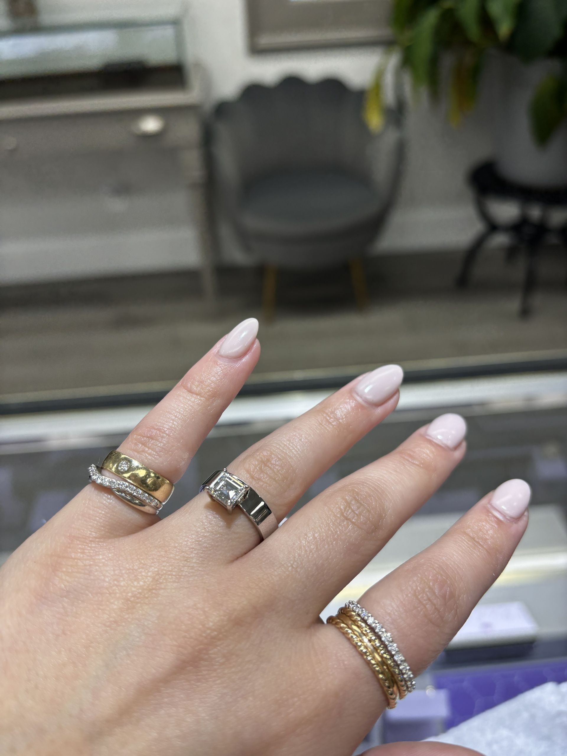 Hand with three rings and pale pink nails, held up in front of a blurred indoor chair.