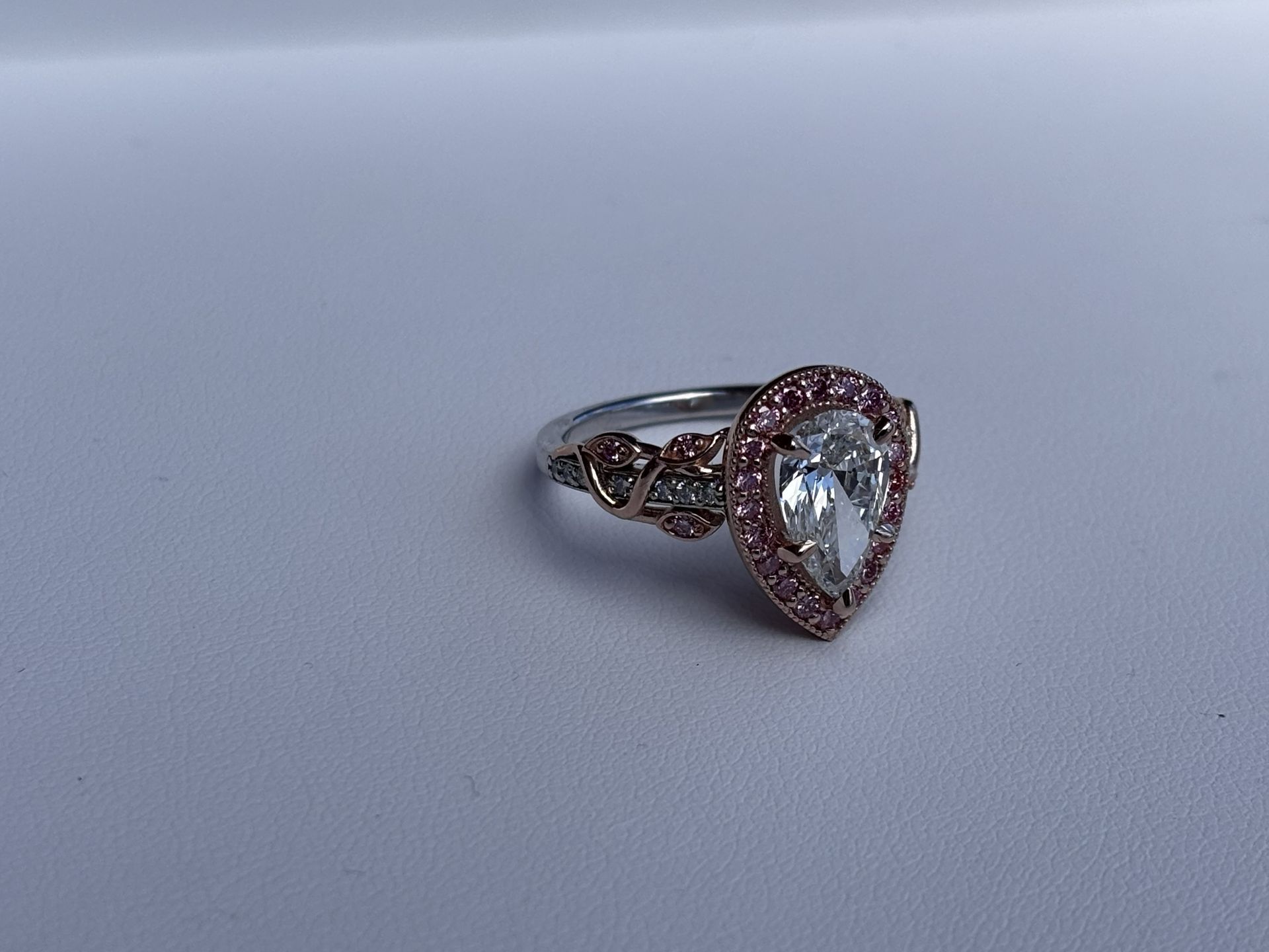 Teardrop-shaped ring with a clear gemstone and pink halo on a light surface