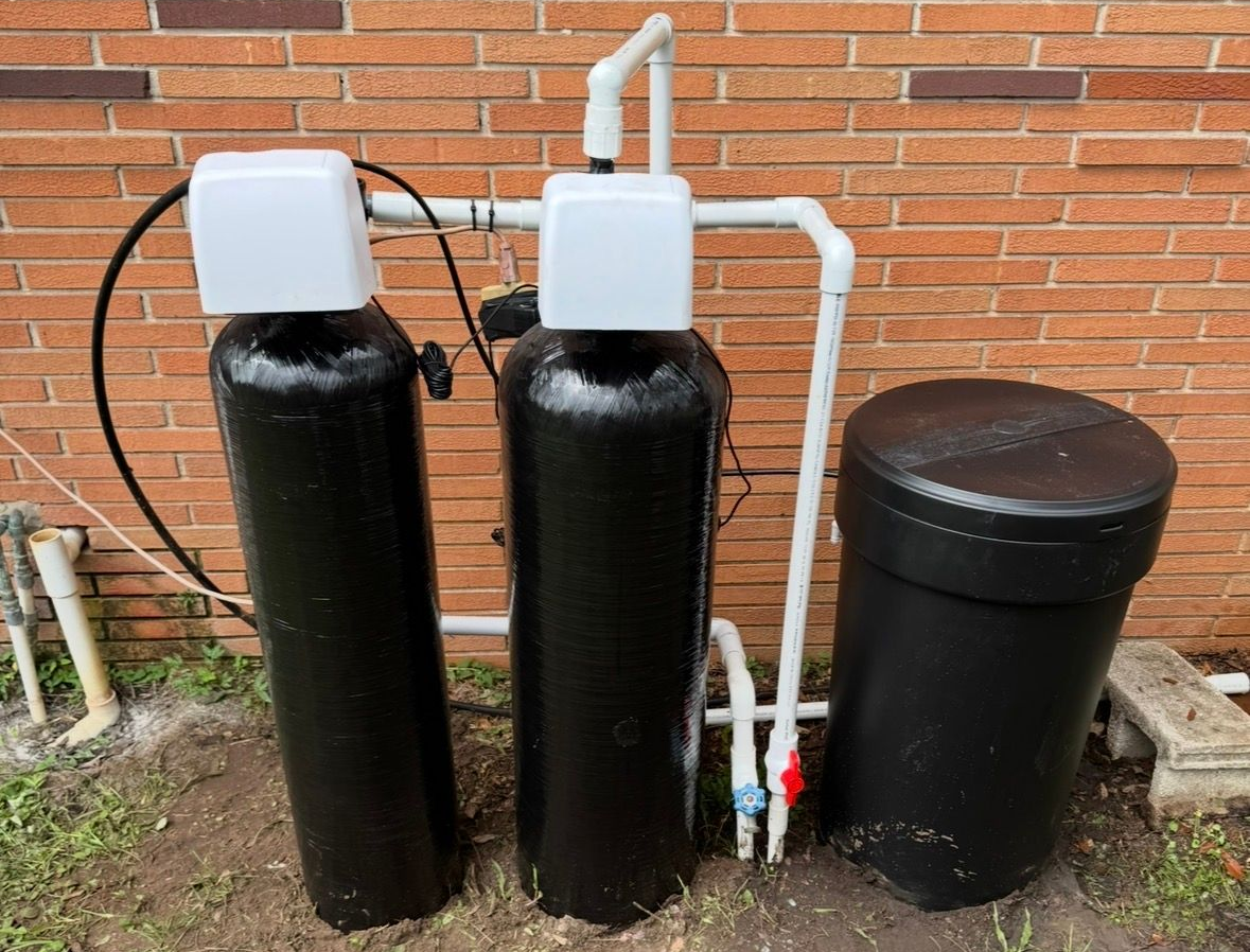 Three black water treatment tanks with white lids and pipes beside a brick wall.
