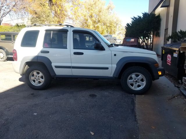 White Jeep Liberty parked next to a building and trash receptacle, on an asphalt surface.