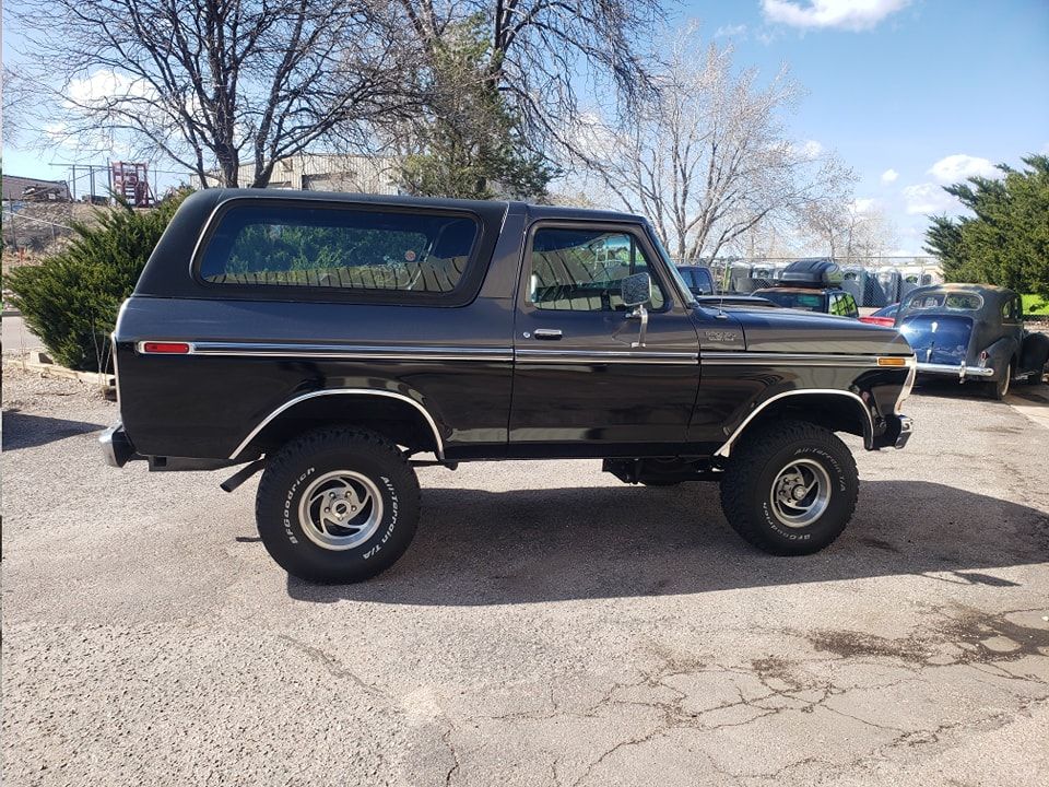 Black and gray Ford Bronco, parked on a paved lot. White-lettered tires and chrome wheels, trees in the background.