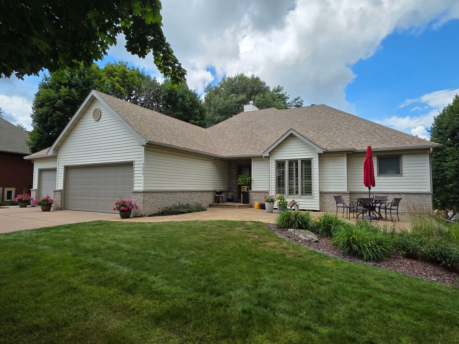 A light-colored, one-story house with a brown roof and green lawn. Patio with a red umbrella and outdoor furniture.