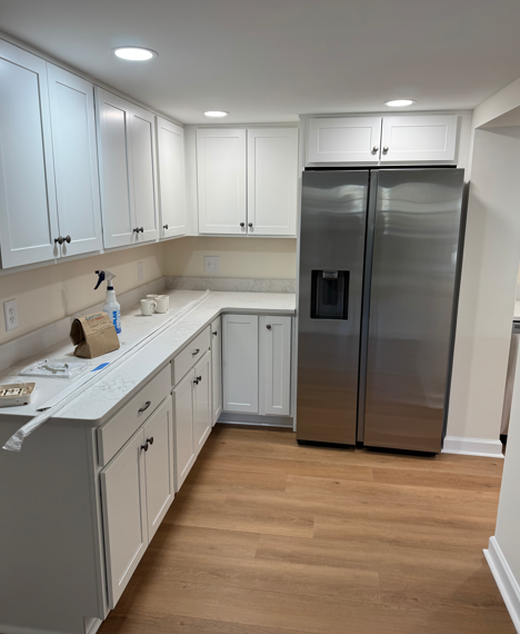 Bright white kitchen with stainless steel refrigerator, light wood floor, and white cabinets.