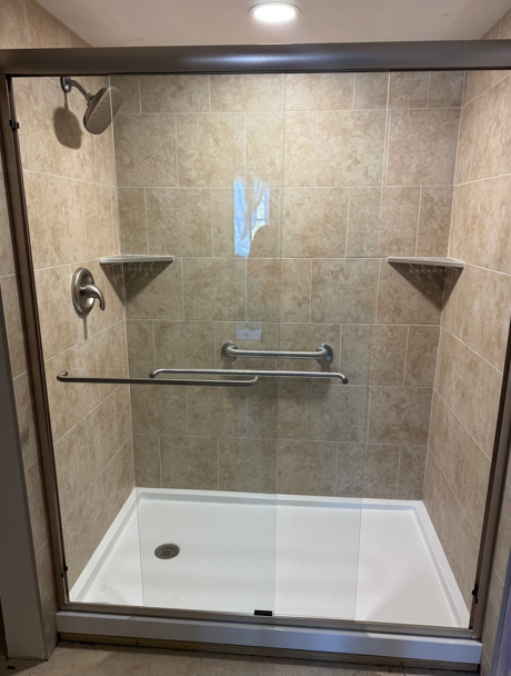 Accessible shower with glass door, grab bar, built-in corner shelves, and tiled walls