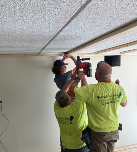Three workers in neon shirts install a ceiling fixture with a power drill indoors.