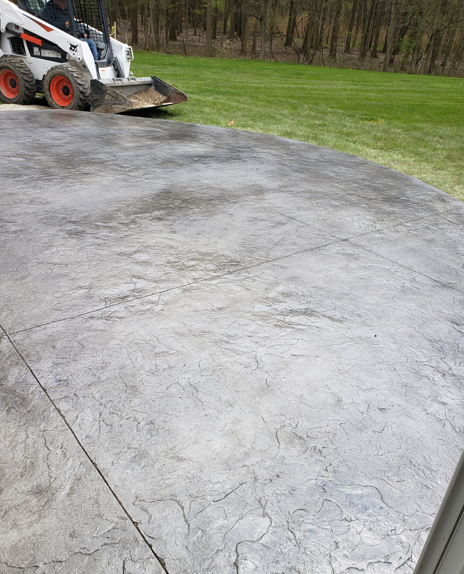 Skid steer smoothing a wet concrete driveway beside a grassy yard and trees