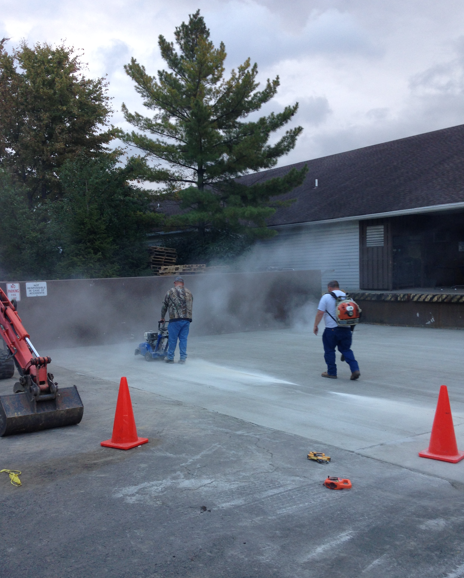 Workers spray sealant on a concrete lot near orange cones as smoke or mist rises beside a building.