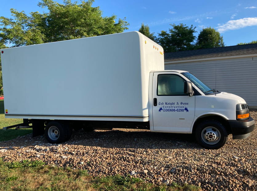 White box truck parked on gravel beside a white building, with trees in the background