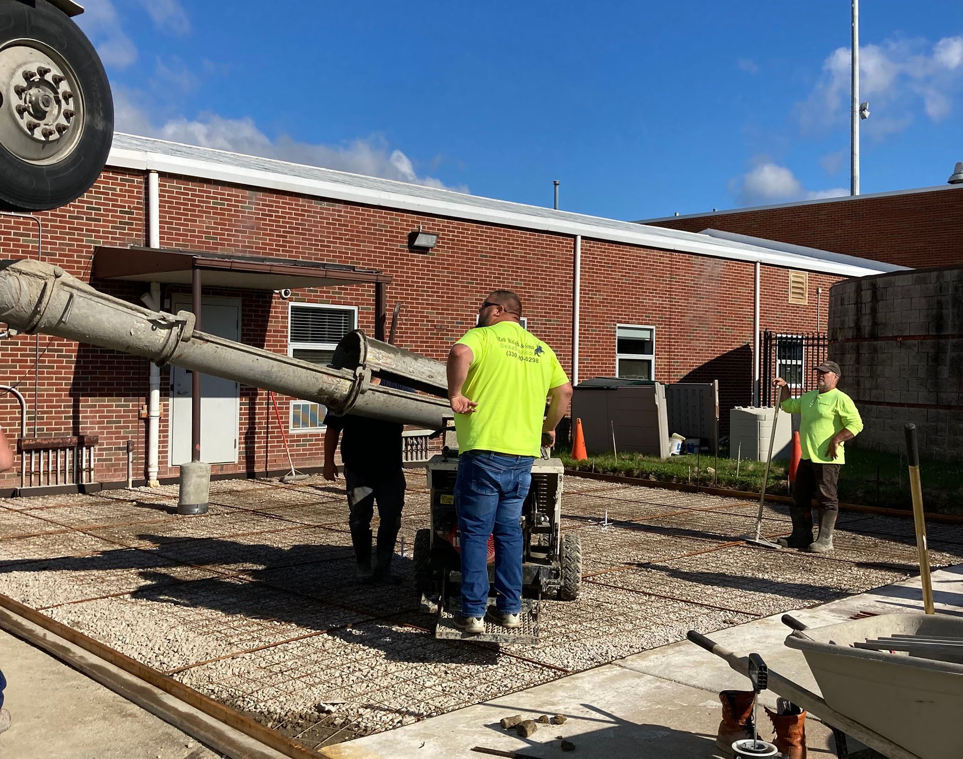 Workers in neon shirts pour concrete at a construction site beside a brick building.