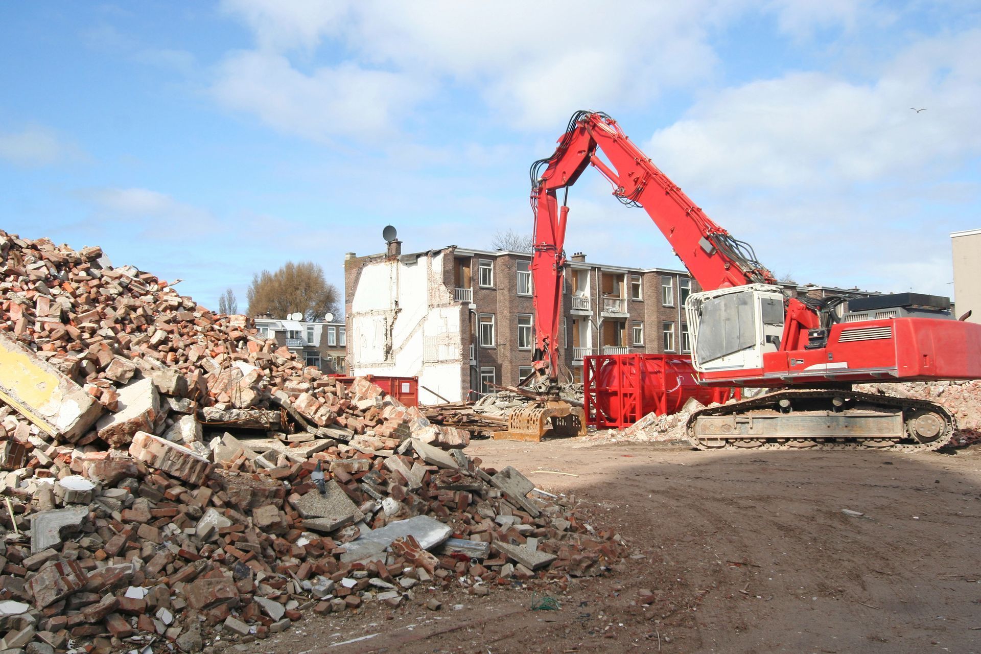 Red excavator demolishing a building beside a large rubble pile under a blue sky