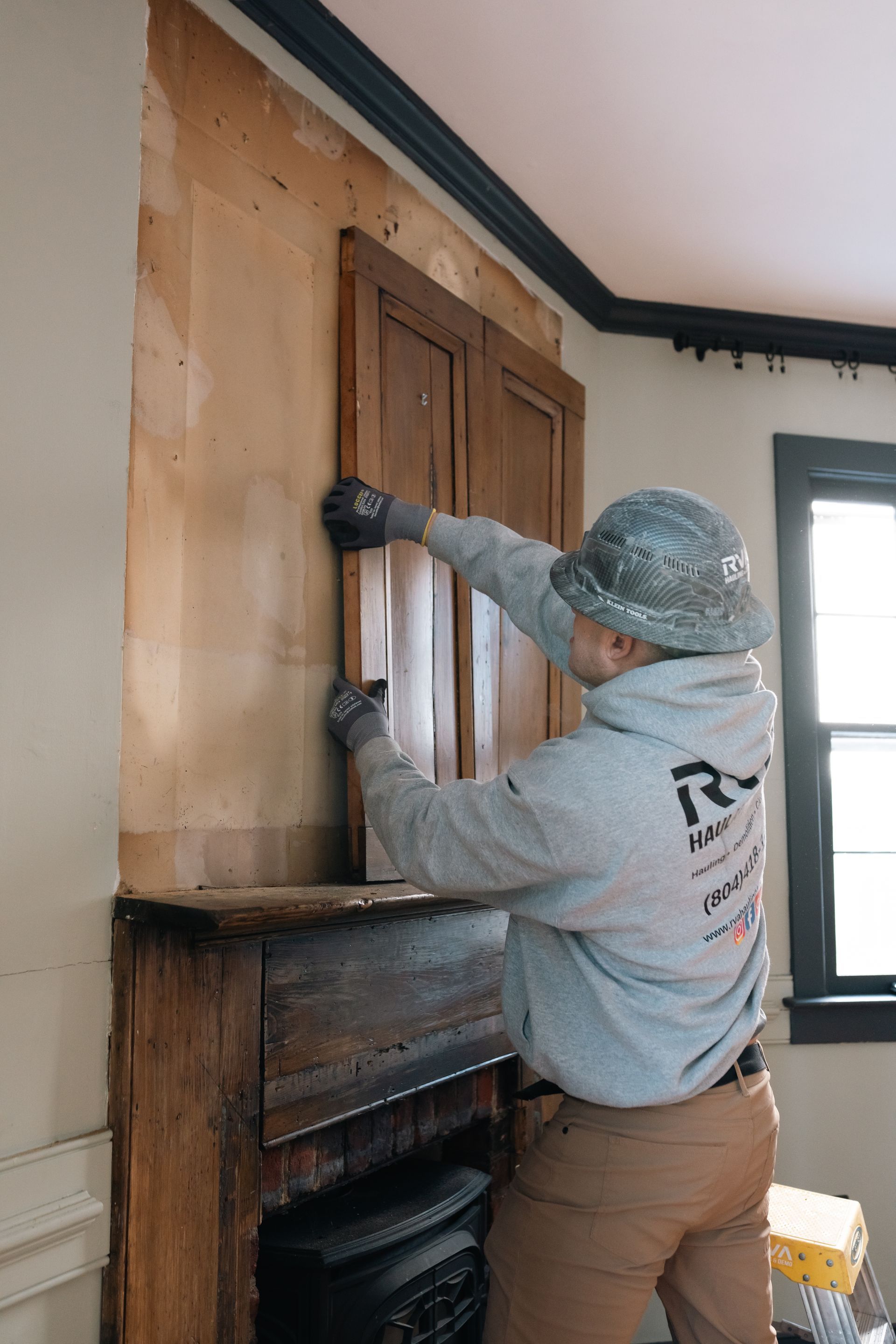 Worker sanding a wooden wall panel beside a fireplace, wearing a hat and gray hoodie.