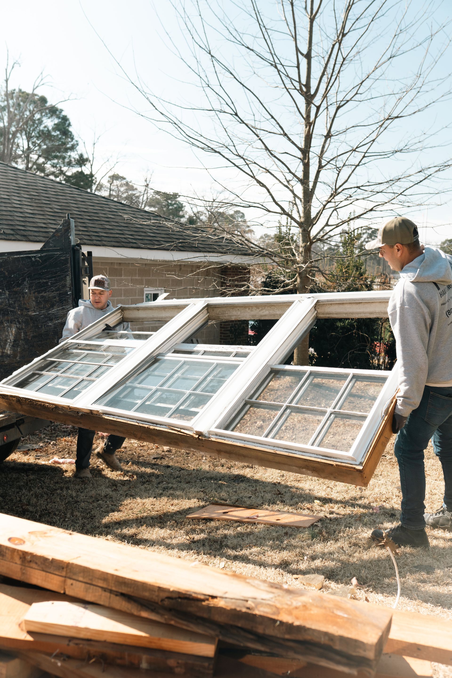 Two men carrying a large white-framed window outdoors near a house and tree.