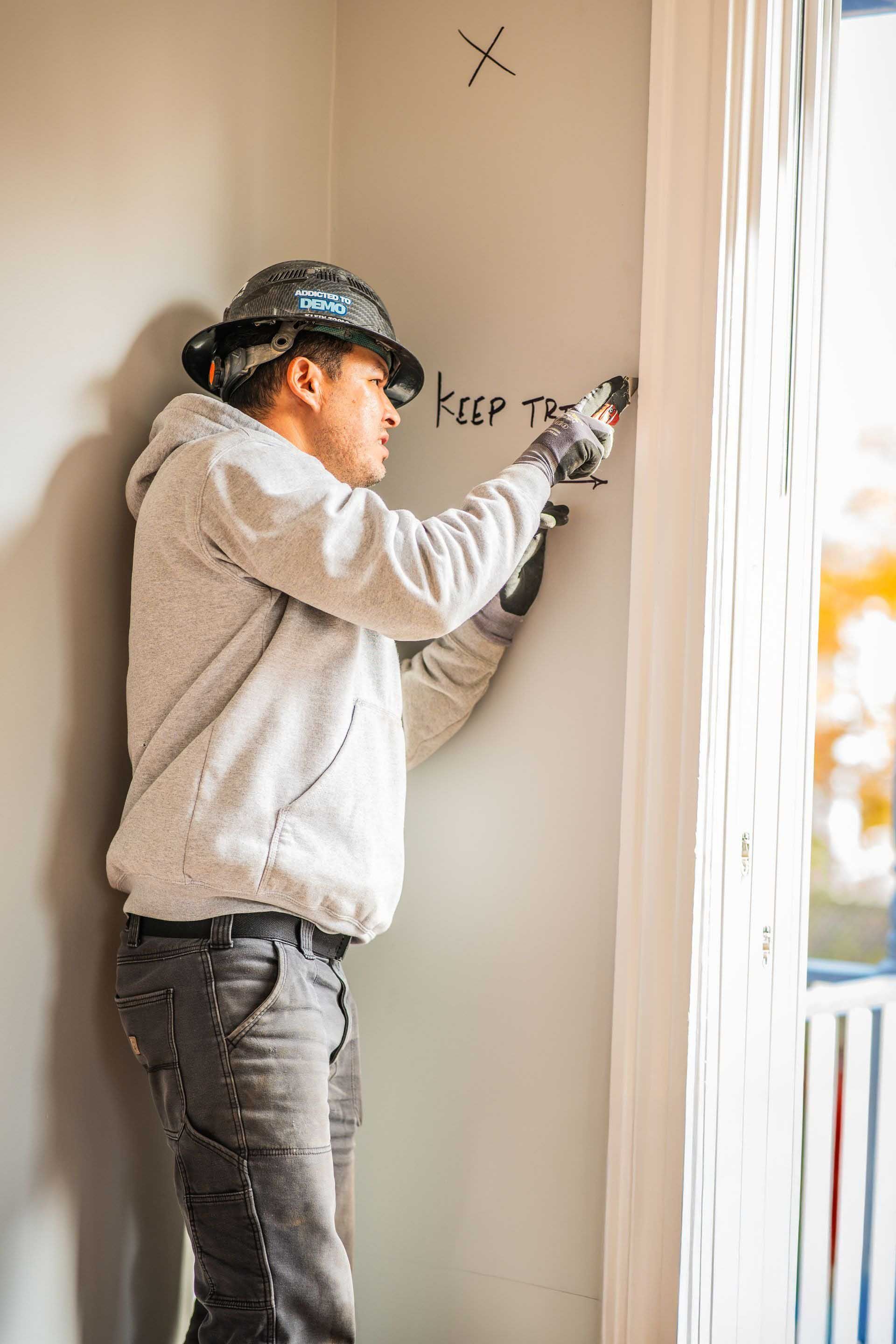 Person painting a wall beside a window, wearing a cap and light hoodie in a bright room