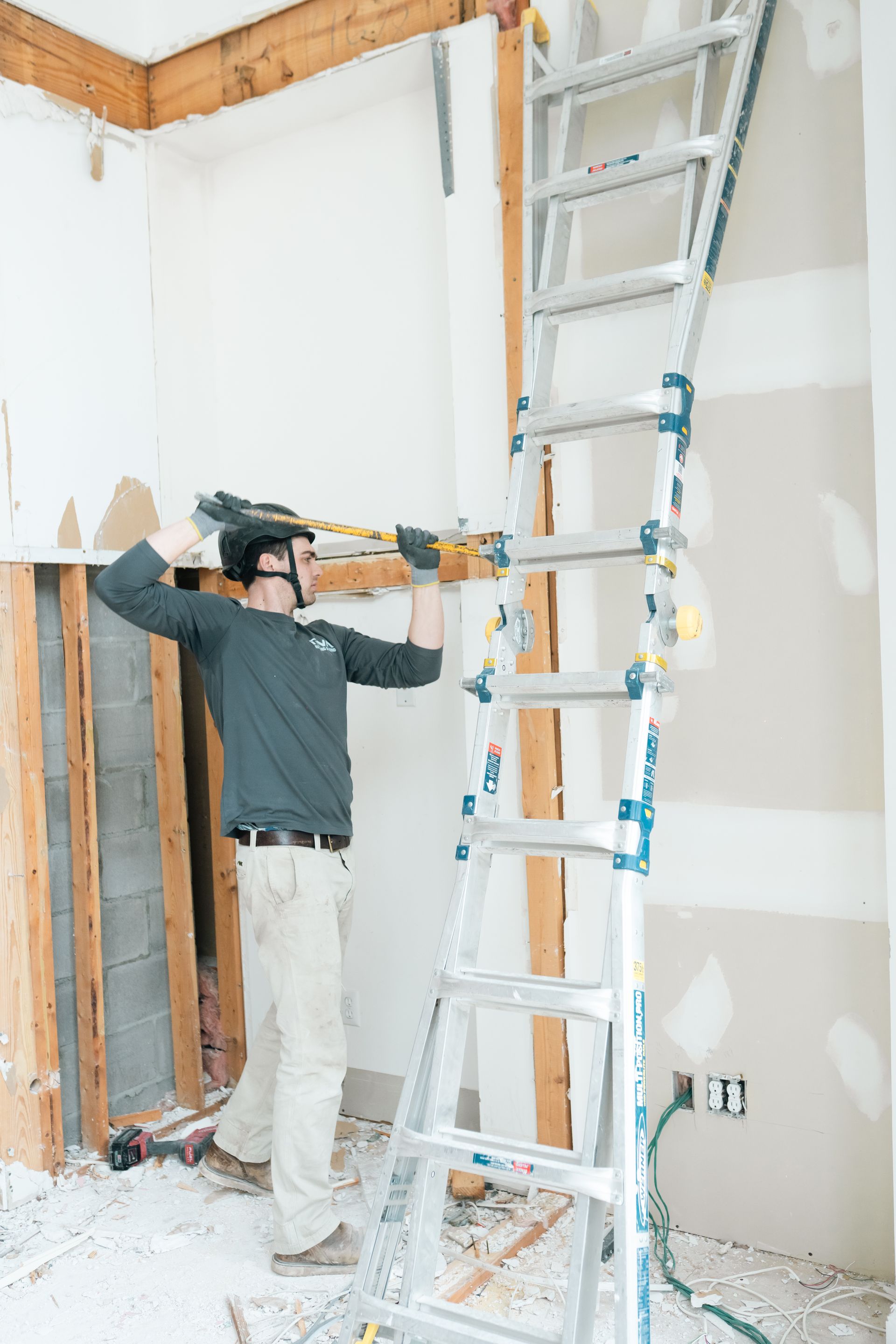 Worker standing on a ladder in a room under construction, with exposed walls and drywall mud patches.