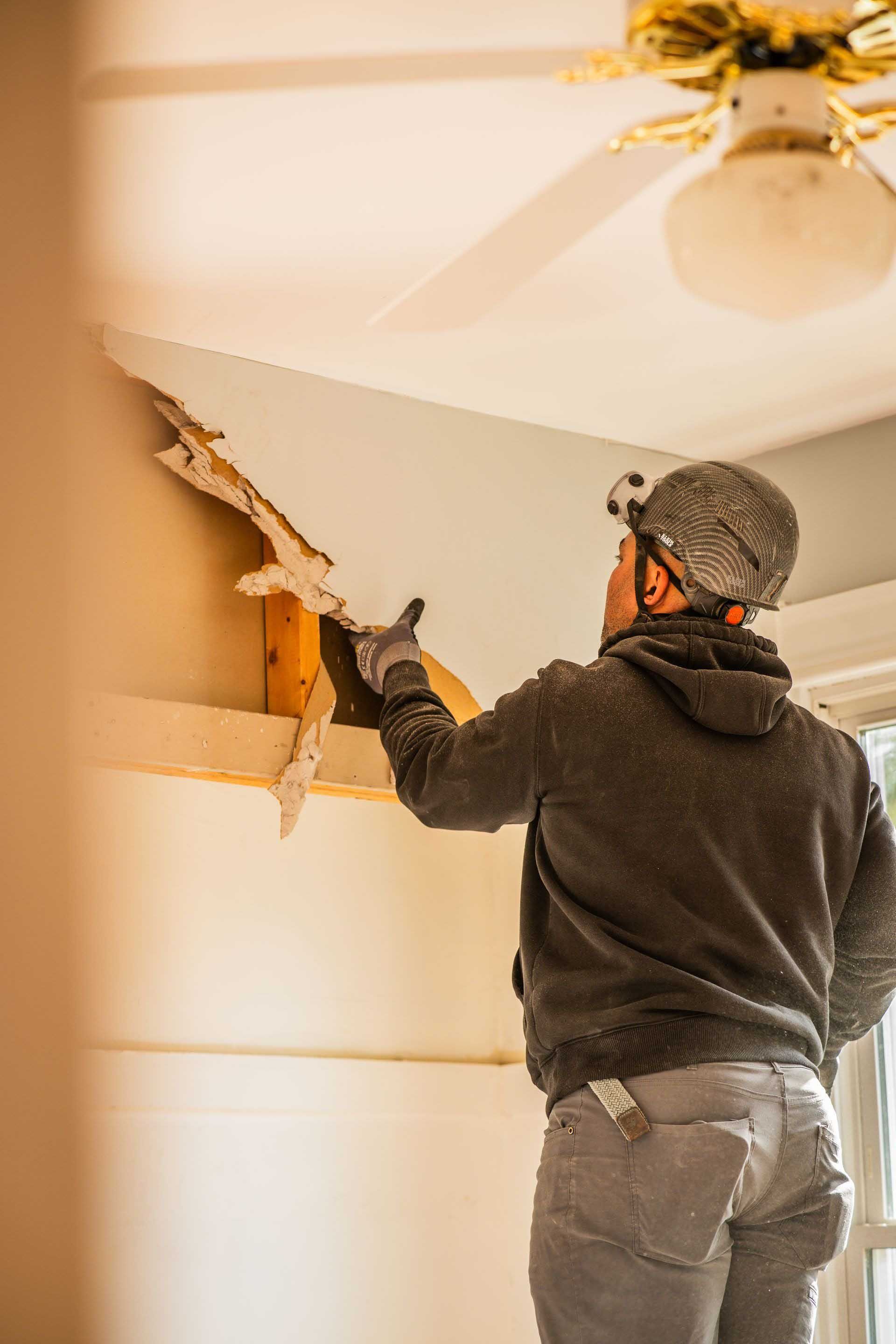 Person using a putty knife to repair drywall damage near a ceiling fan in a room